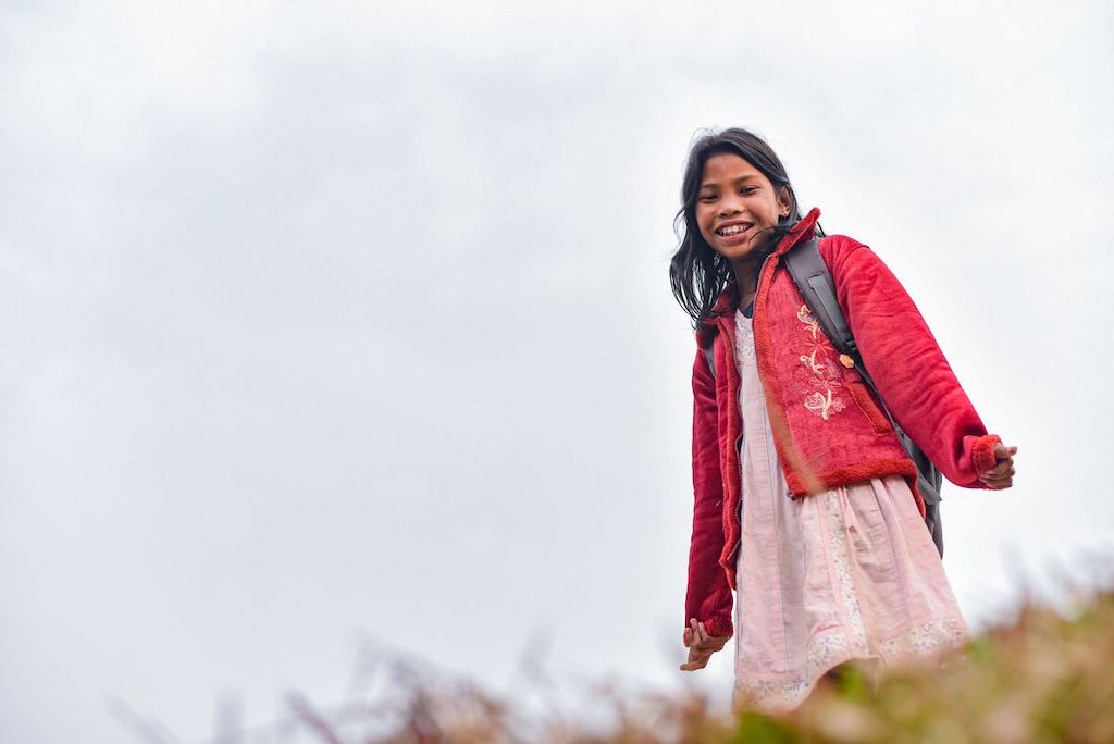 Anushka stands on a hill wearing a pink dress, red coat and her school backpack.