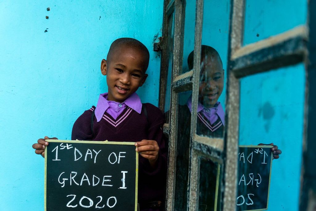 Hamisi stands against a blue wall holding a chalkboard that says "1st day of grade 1, 2020".