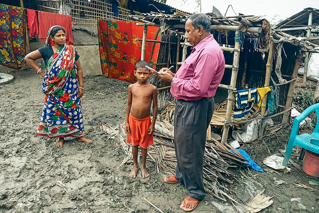 A Compassion centre director stands with a mom and her son in front of their home, taking notes on a notepad as he speaks to them.