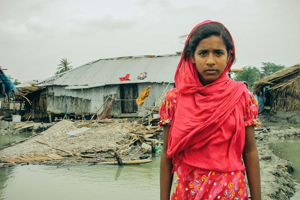 Aduri, wearing a red dress and headscarf, stands in front of her collapsed bamboo home.