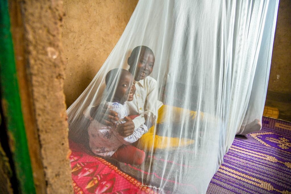 Two little boys sit on a bed under a mosquito net. They're smiling.