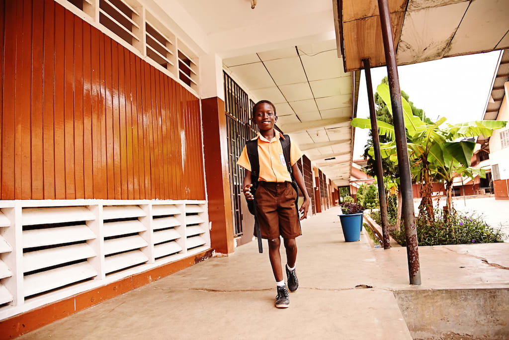 A boy walks on a path outside his school, wearing a uniform and backpack.