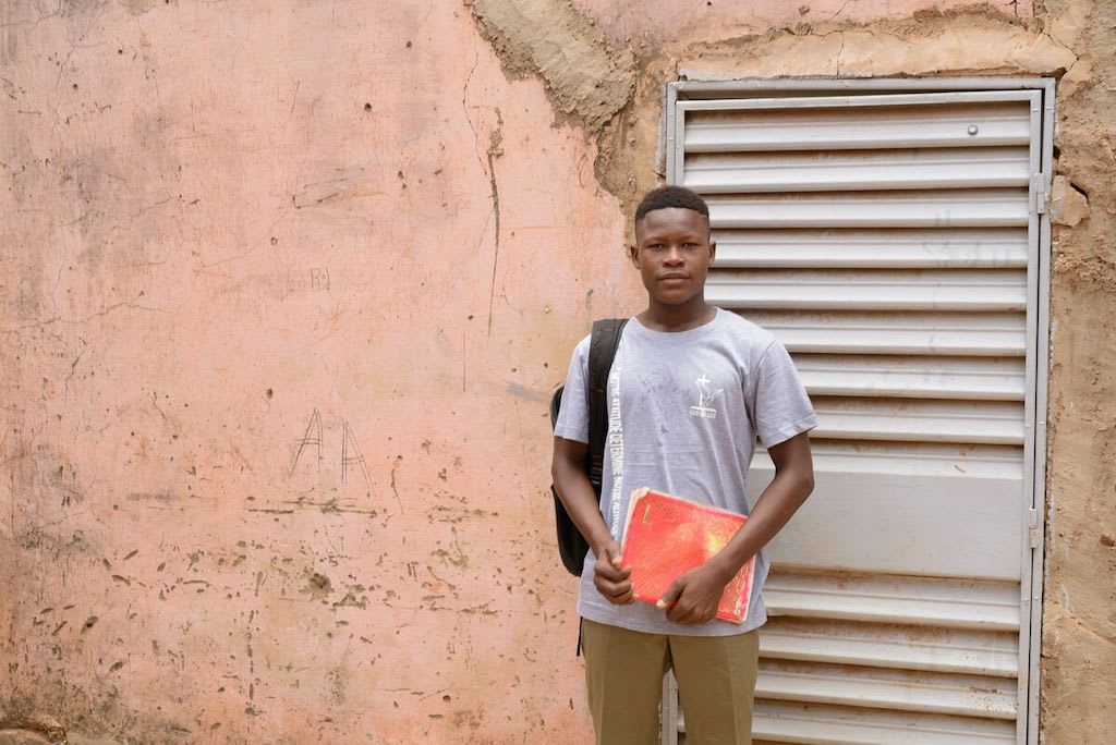 Abdoul stands boy a door, wearing a backpack and holding a red notebook.