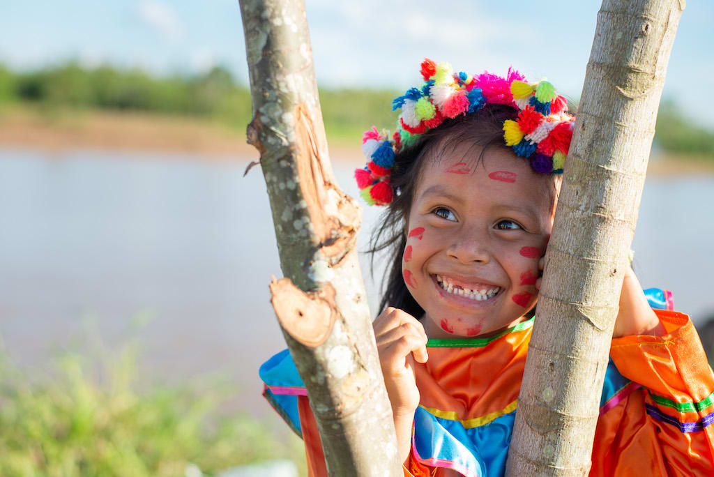 A young Shawi girl wearing a colourful headpeice and dress smiles and looks away from the camera.