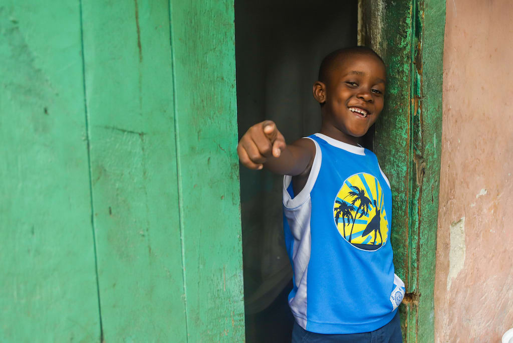A boy in a blue tshirt stands in a green doorway smiling and pointing a finger in a pose.