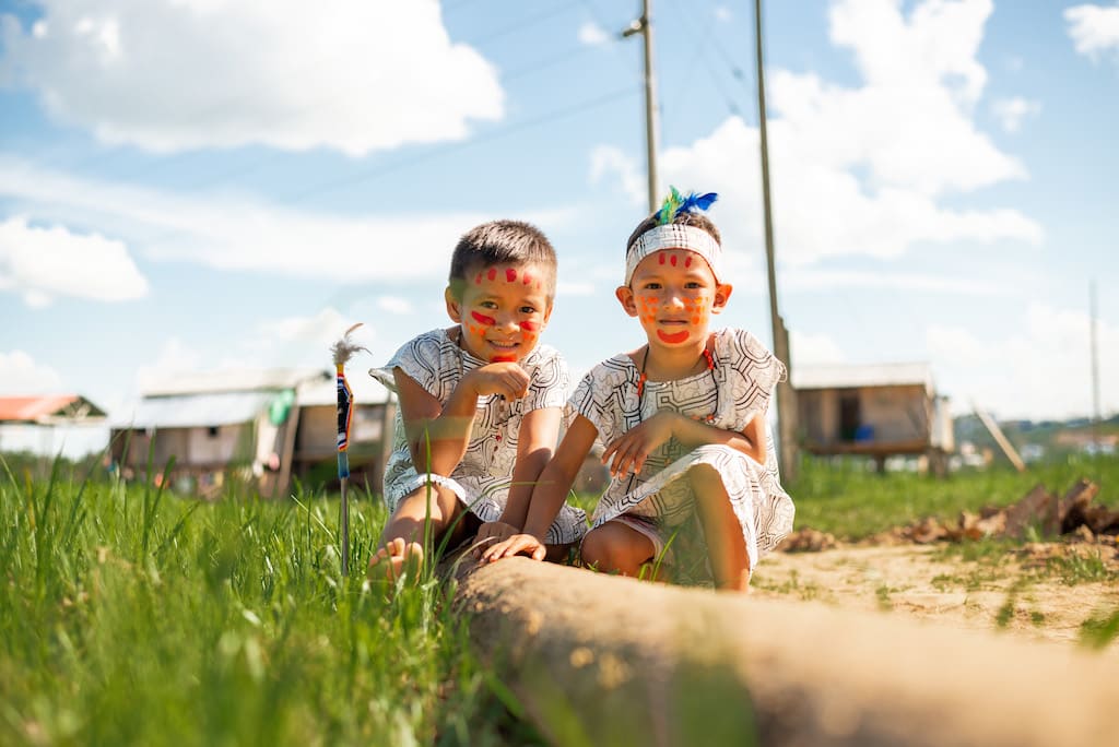 Two boys sit crouched in a field, wearing their tribe's traditional clothing.