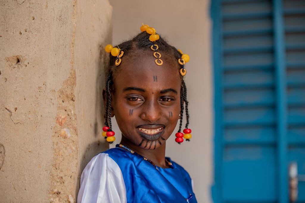 Oureira is wearing a blue and white traditional dress with red and yellow beads in her hair. She is also wearing black makeup on her face.