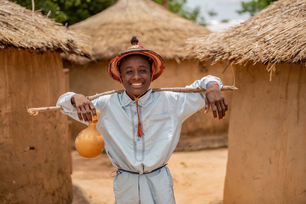 Abdoul is wearing yellow pants and a long light blue shirt with a hat. He is carrying a stick and a calabash cup. He is standing in front of three huts in his community.