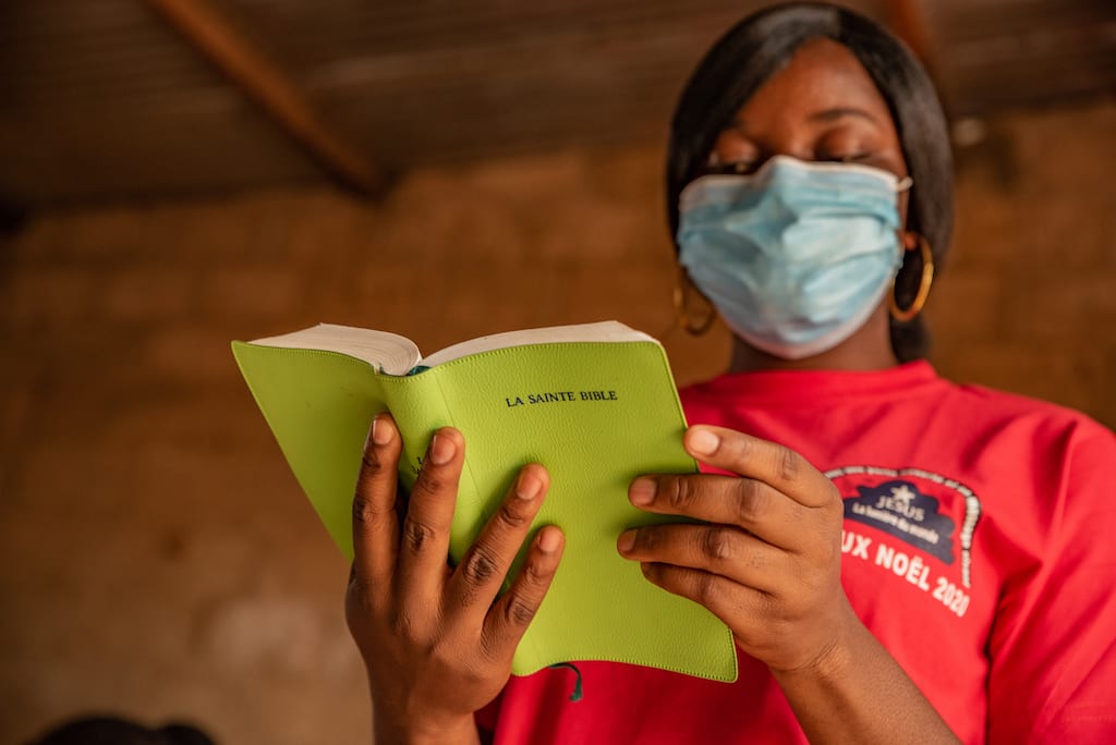 Rainatou is wearing a red shirt and a face mask. She is inside the center and is reading her green Bible.