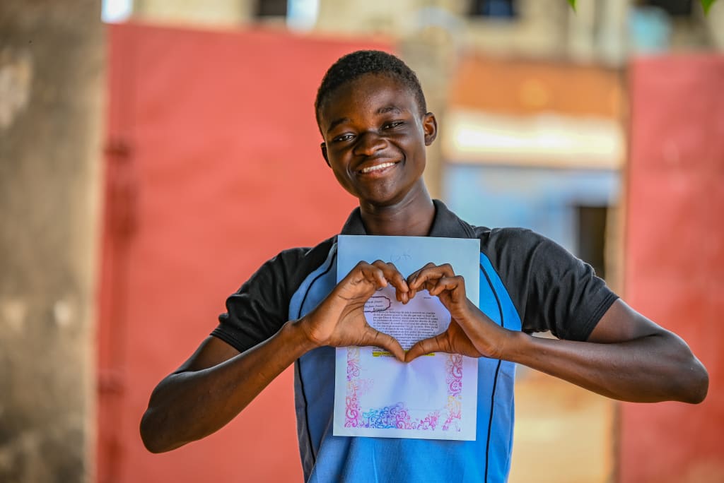 A teenage boy in a blue and black tshirt holds his hands in a heart shape in front of a letter