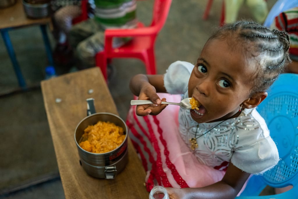 a young girl takes a mouthful of stew or curry from a small tin can. She experiences food insecurity and would be at risk for malnutrition if not for the support from the Compassion centre.