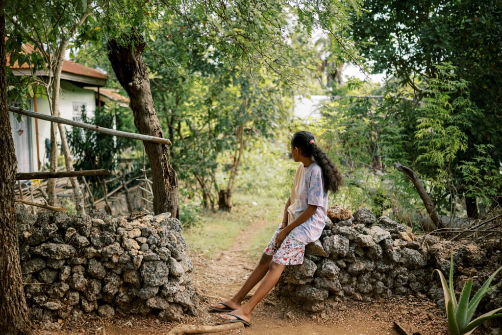 A girl with long brown hair wears white and sits on a stone wall looking towards the trees.