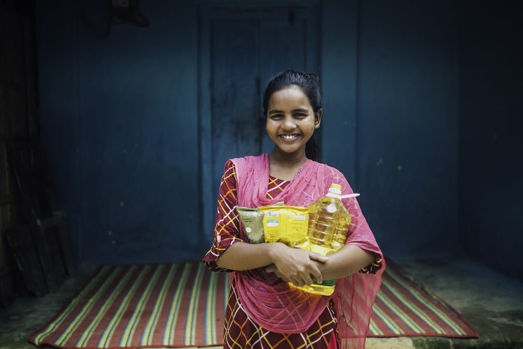 Girl in a pink Sari smiles at the camera while holding rice and oil. The background is dark blue.