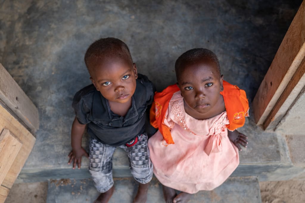 Two young children with dark skin and sad faces sit on the step of their home. Nikoa and Emiliana were facing malnutrition before food support was given.