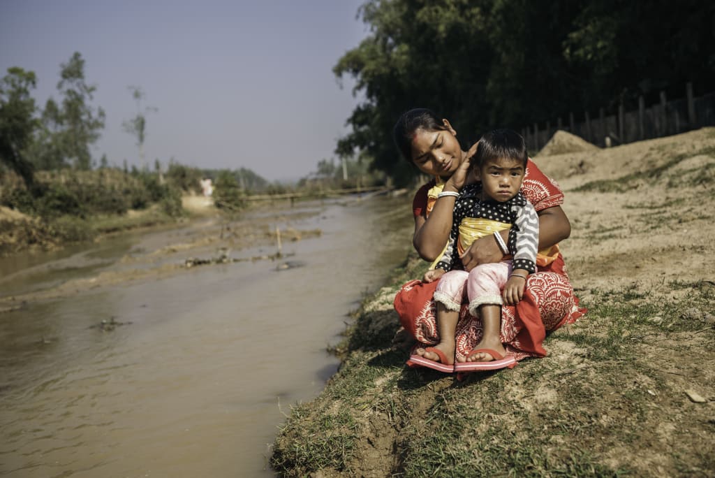A woman in red crouches down and holds a toddler by a riverbank.