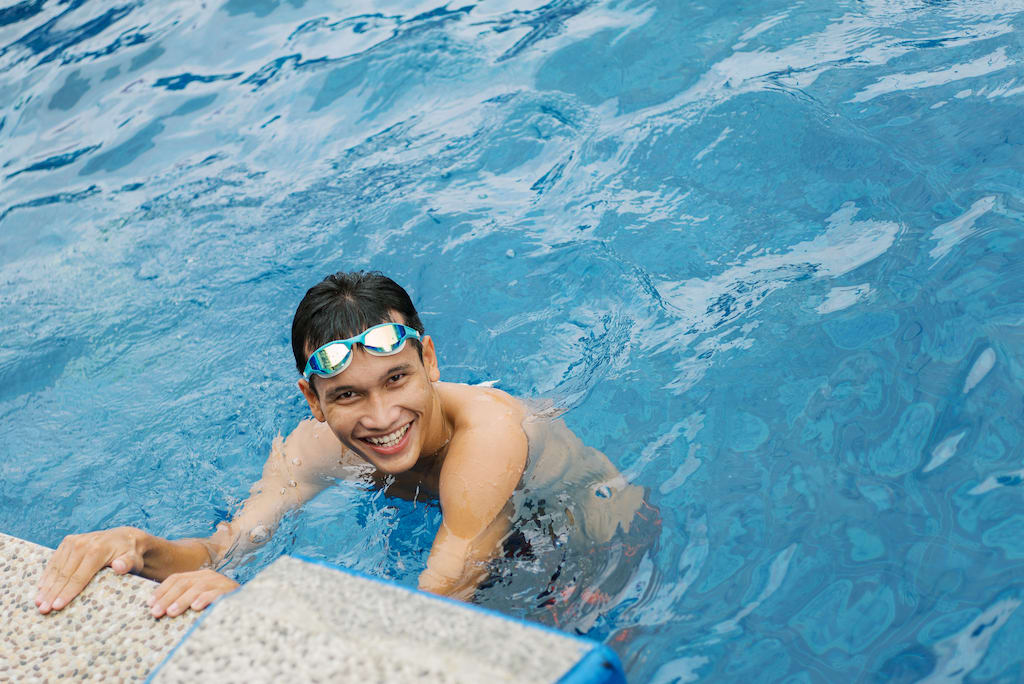 A young man is seen in the pool smiling with goggles on his head.