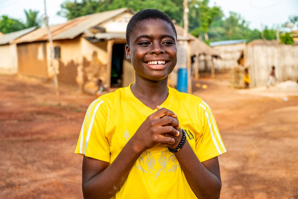 A teenage girl in a yellow tshirt stands smiling with her hands clasped.