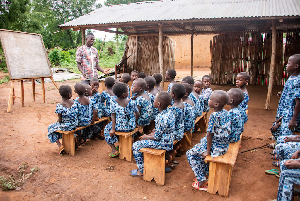 A group of students in Togo, all wearing blue uniforms, sit on benches in an outdoor courtyard, listening to a workshop facilitator.