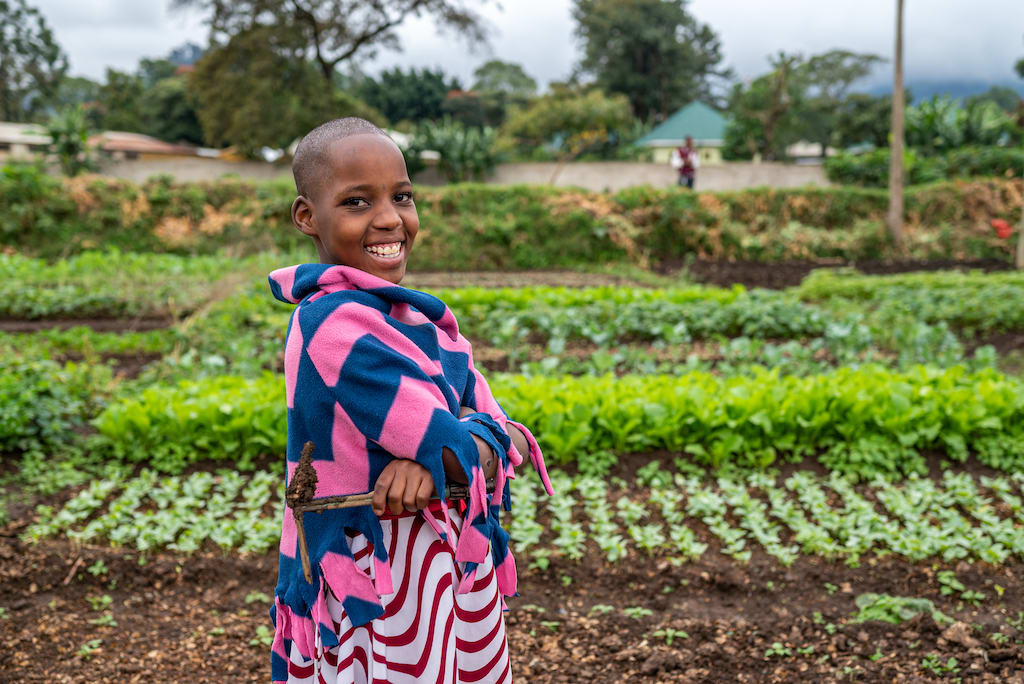 Hosiana, in a pink and blue poncho, is standing in her grandmother’s vegetable garden with her arms crossed in front of her.