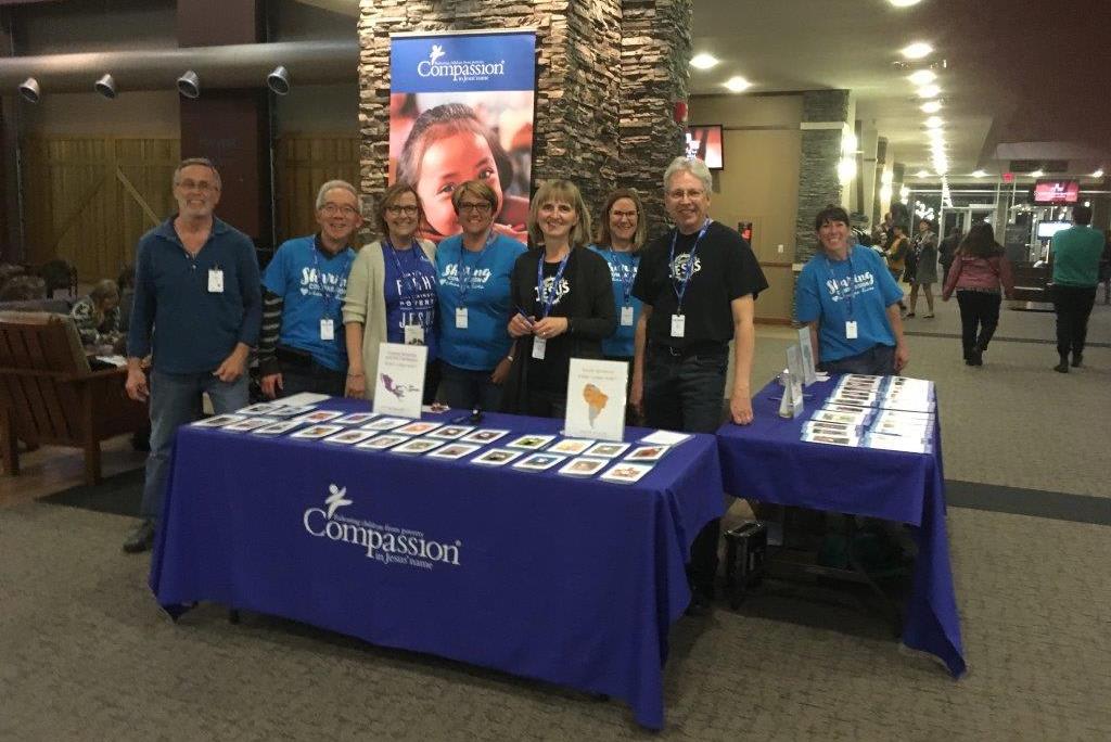 A group of volunteers stand at a Compassion display set up at an event promoting Compassion and child sponsorship.