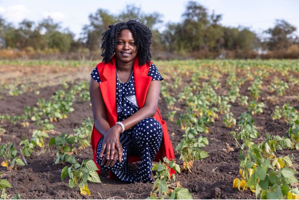 Janet, wearing polka dots and a red vest, crouches down in a field of crops.