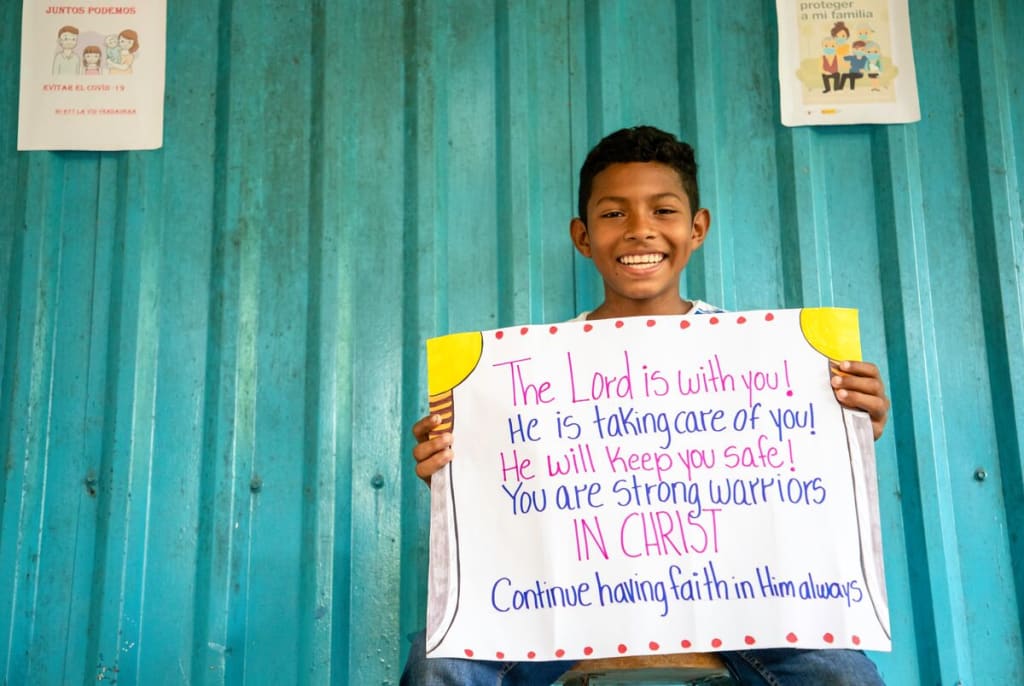 A boy sits against a green wall holding a sign that says, "The Lord is with you! He is taking care of you! He will keep you safe! You are strong warriors in Christ. Continue having faith in Him always."