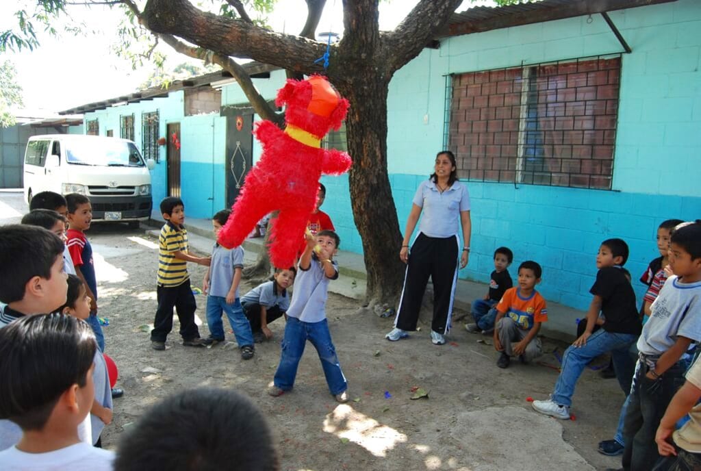 Little boy hits a red pinata surrounded in friends, outside of a blue painted building