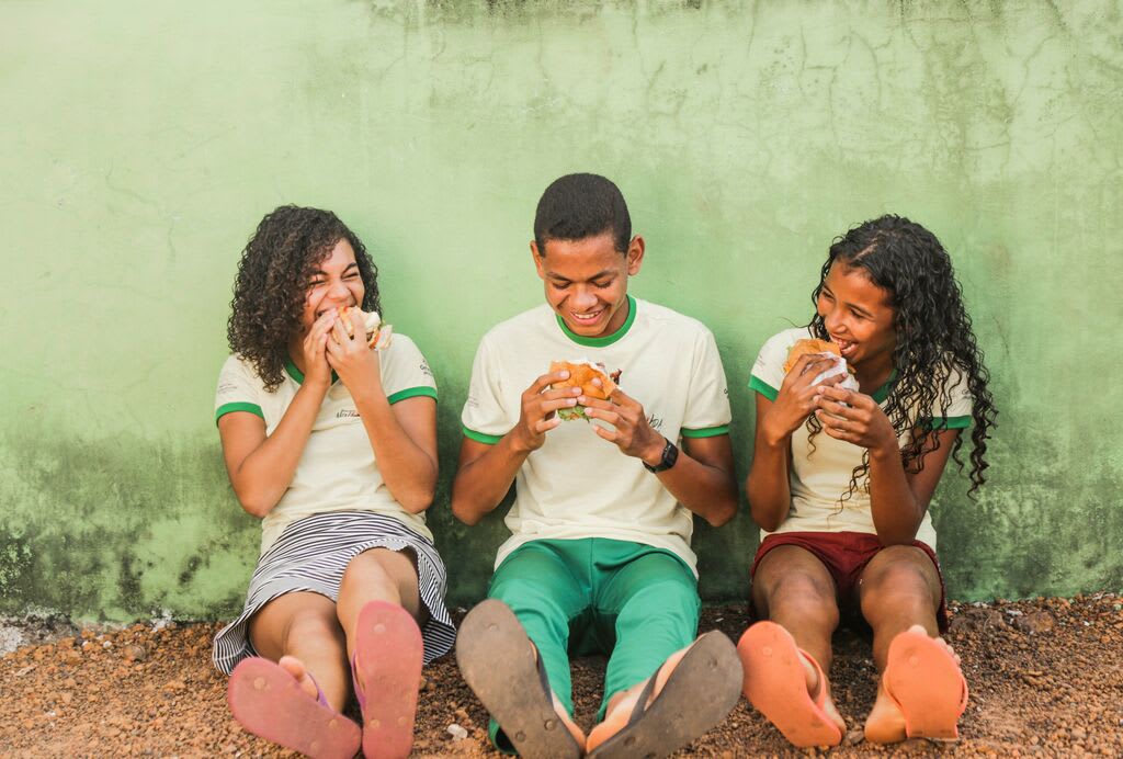 Three teens smile while eating a hamburger each. They are lsitting against a green wall.