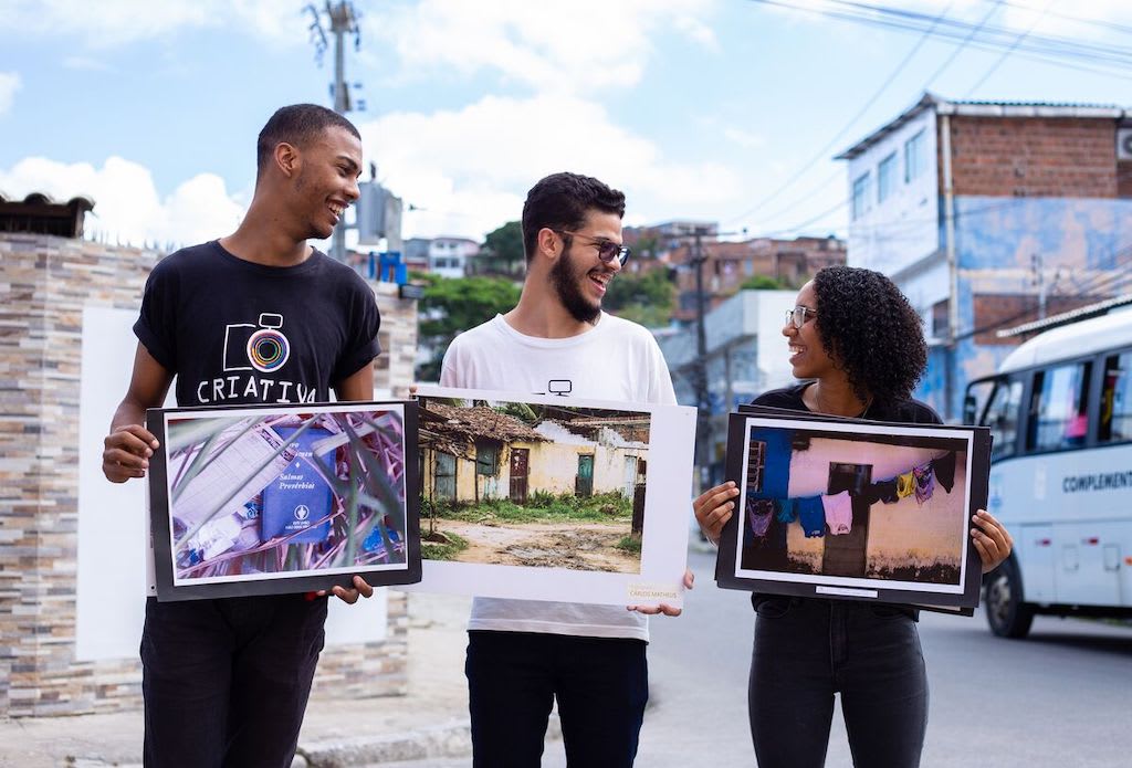 A group of three students pose in a street, each holding a photograph that they took.