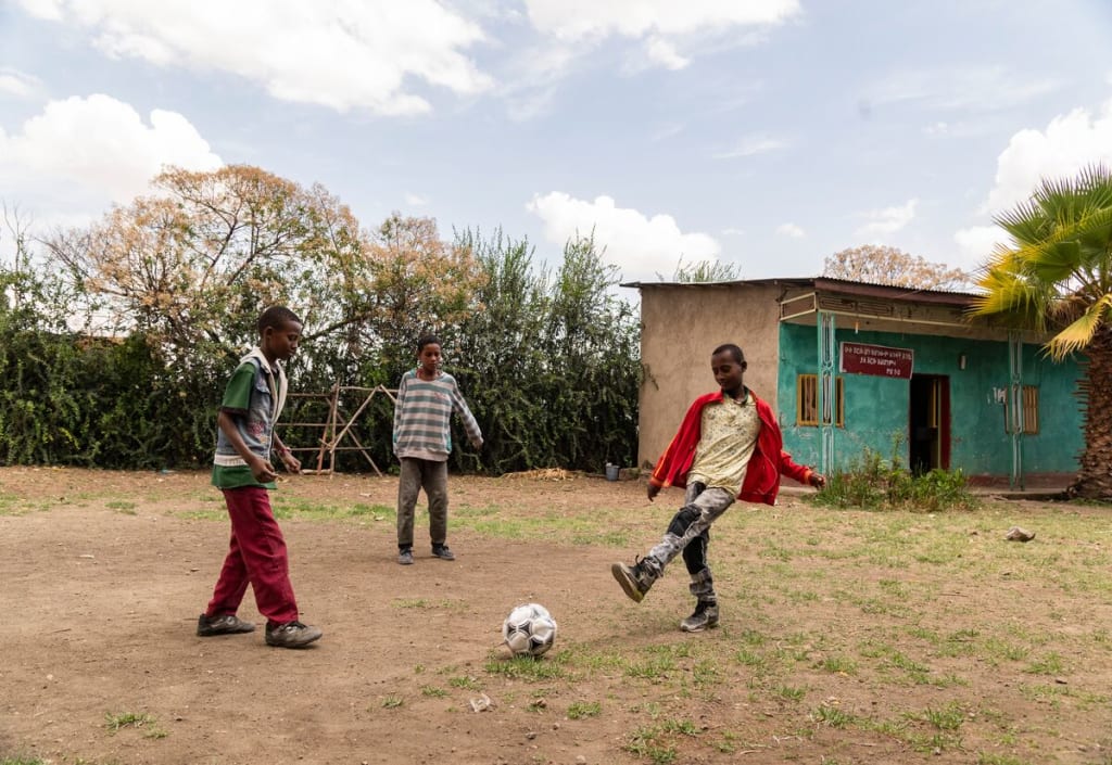 Abel, in a red jacket, is outside in the yard at the church kicking around a soccer ball with friends. There is a tan and teal building in the background.