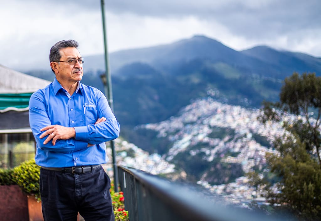 Pastor Sixto Gamboa is wearing a blue shirt. He is standing outside with his arms crossed and is looking out at the mountains in the background.
