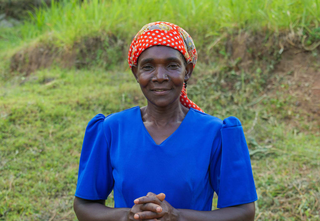 A woman in a blue dress and red head covering smiles at the camera.