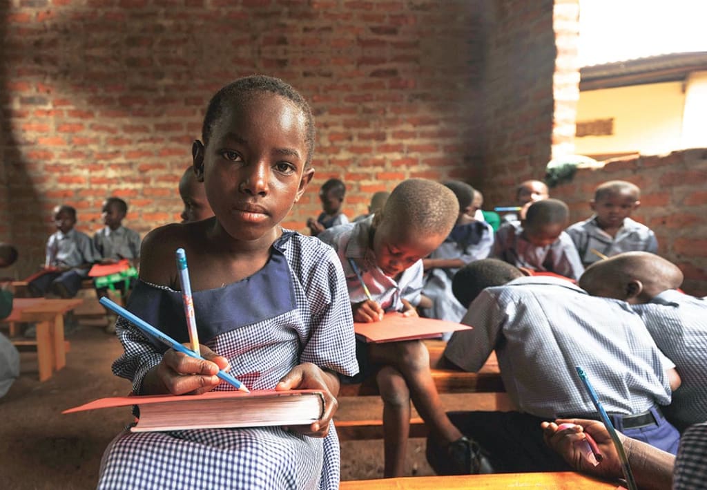 A child sits on a bench in brick classroom. She writes with a pencil, using her textbook as a hard surface to write on.
