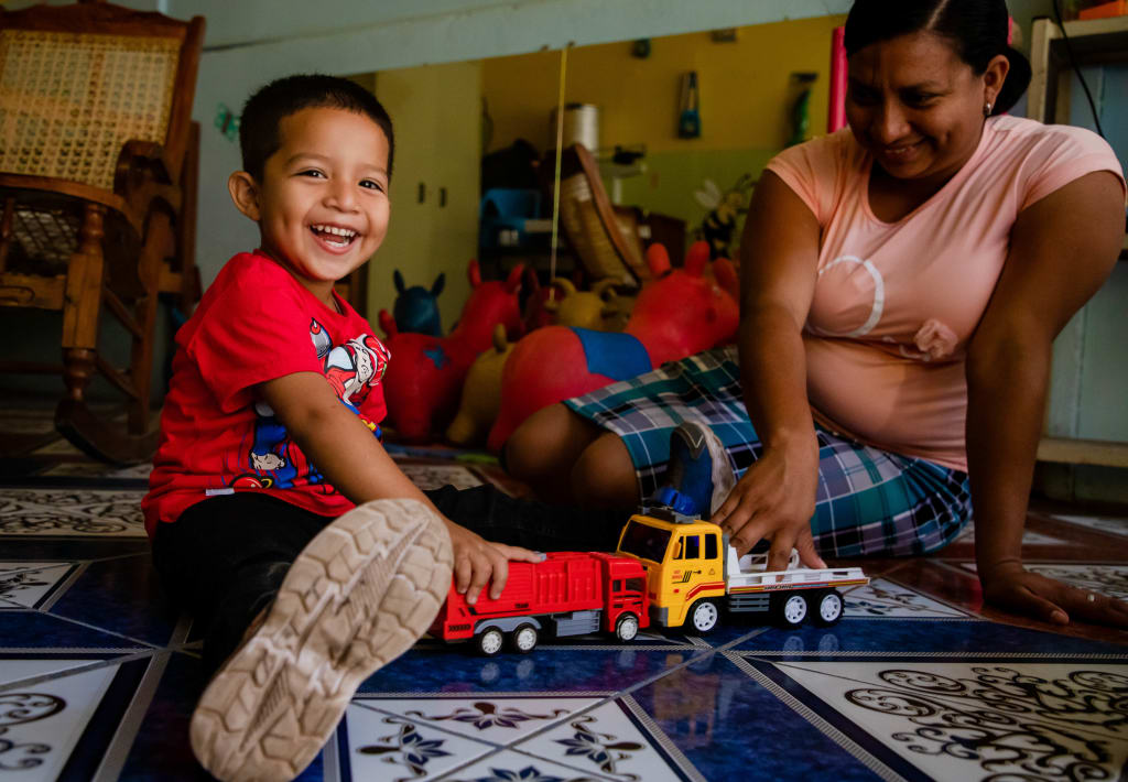 A little boy in red sits on the ground and plays with a red toy truck. A caretaker in pink sits behind him and smiles.