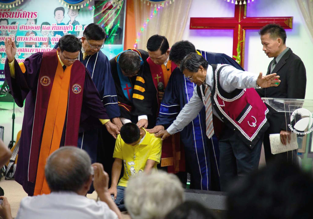 A group of pastors is praying over a boy who is kneeling on the ground. There are colourful lights and a cross in the background.