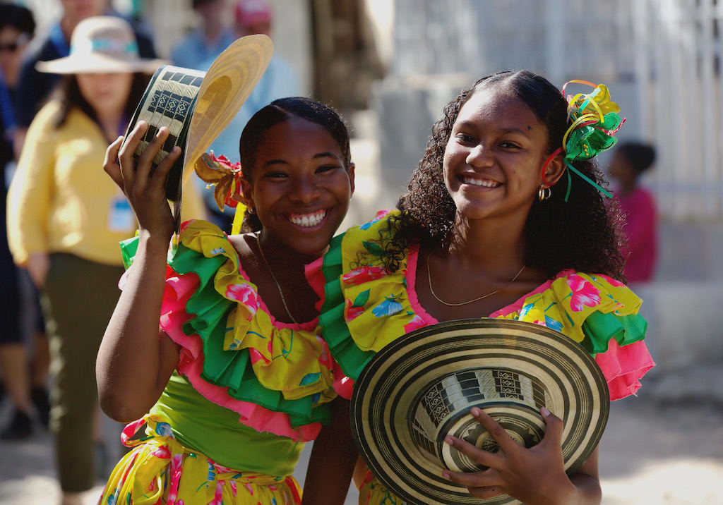 Two young women in Colombia dressed in colourful dresses and holding straw hats.