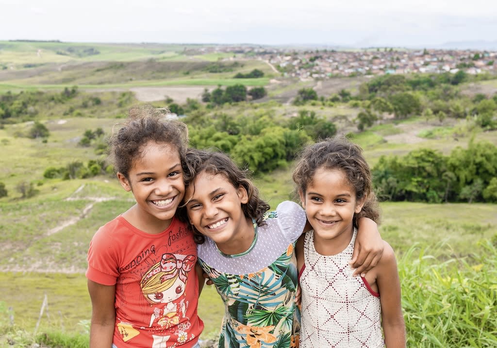 Three girls smiling together on the top of a mountain. One is wearing a red shirt, the middle girl a white and green shirt and the third a yellow shirt.
