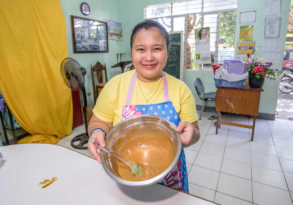 An Asian woman wearing a yellow t-shirt and apron shows a bowl she is stirring.
