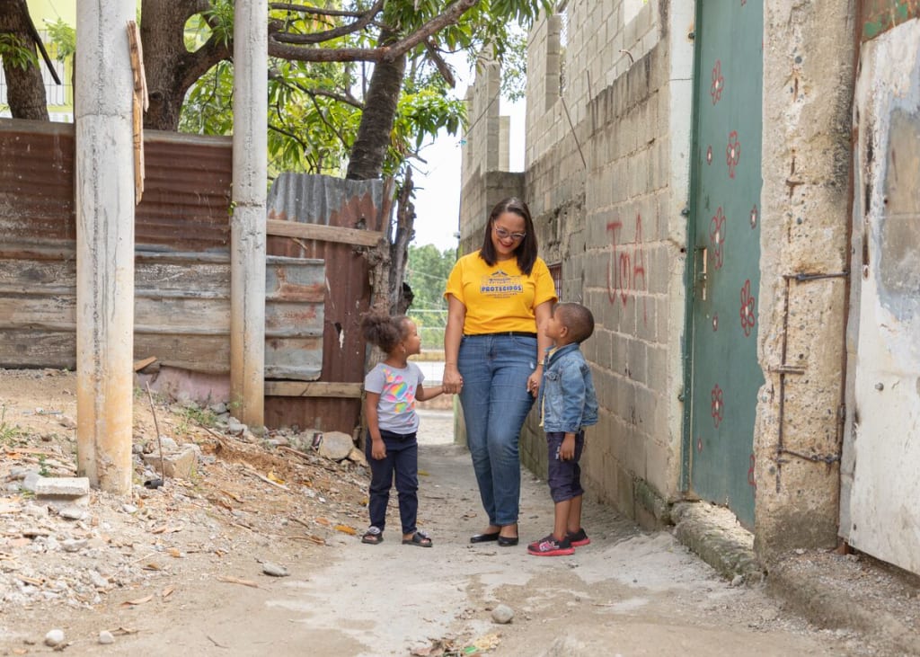 A woman in a yellow shirt stands holding two young children's hands