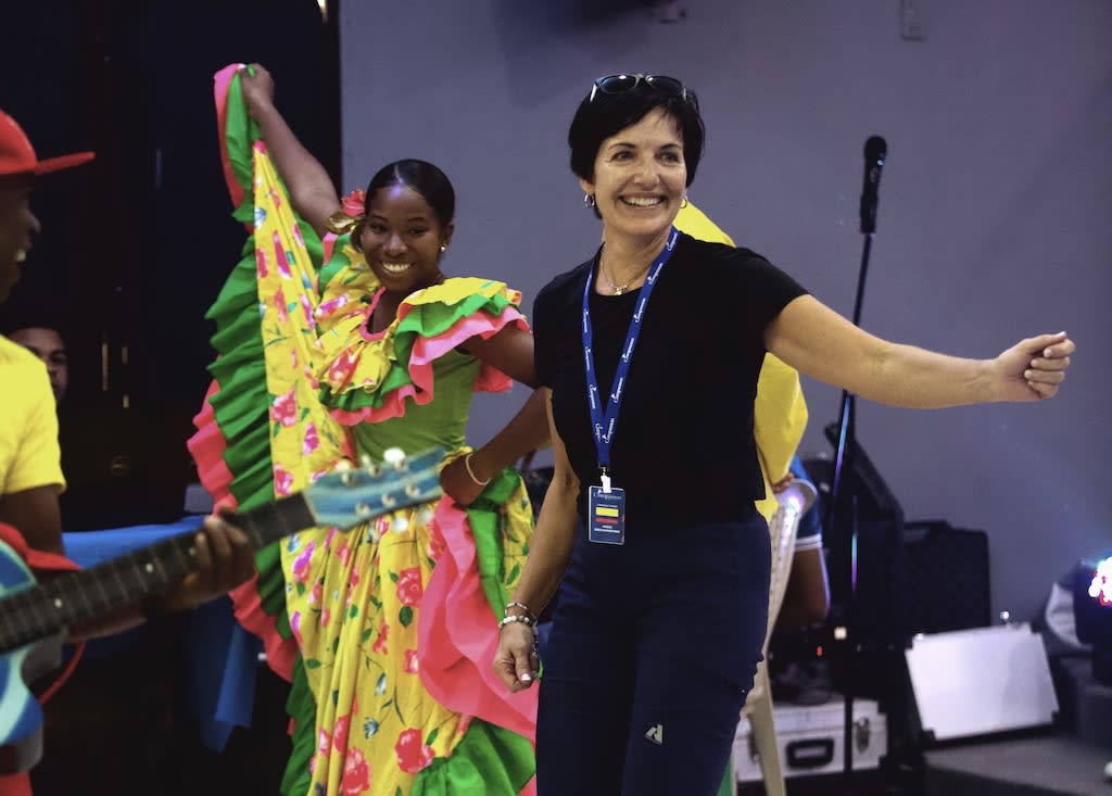 Marie is wearing a black t-shirt and a lanyard, dancing with some youth dressed in colourful dresses at a Compassion centre in Colombia
