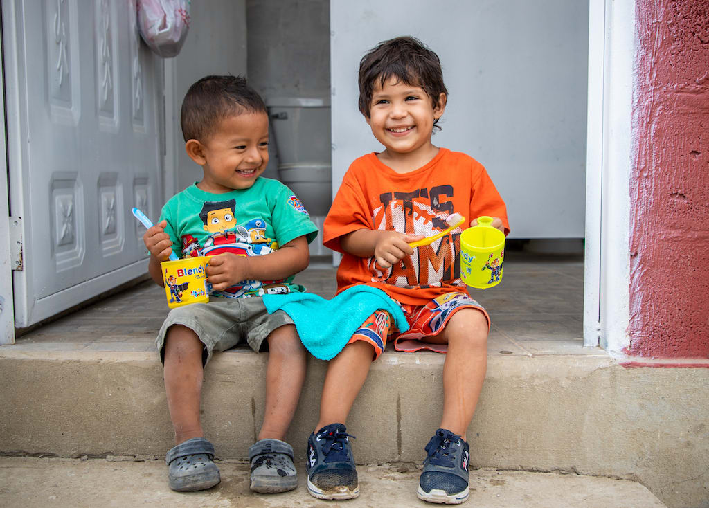 Two boys sit on a curb smiling with toothbrushes in their hands.