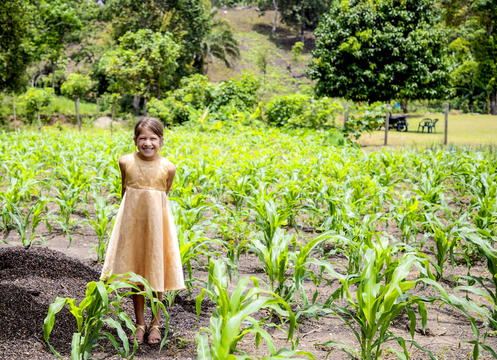 Little girl wearing a yellow dress stands beaming in a green field.