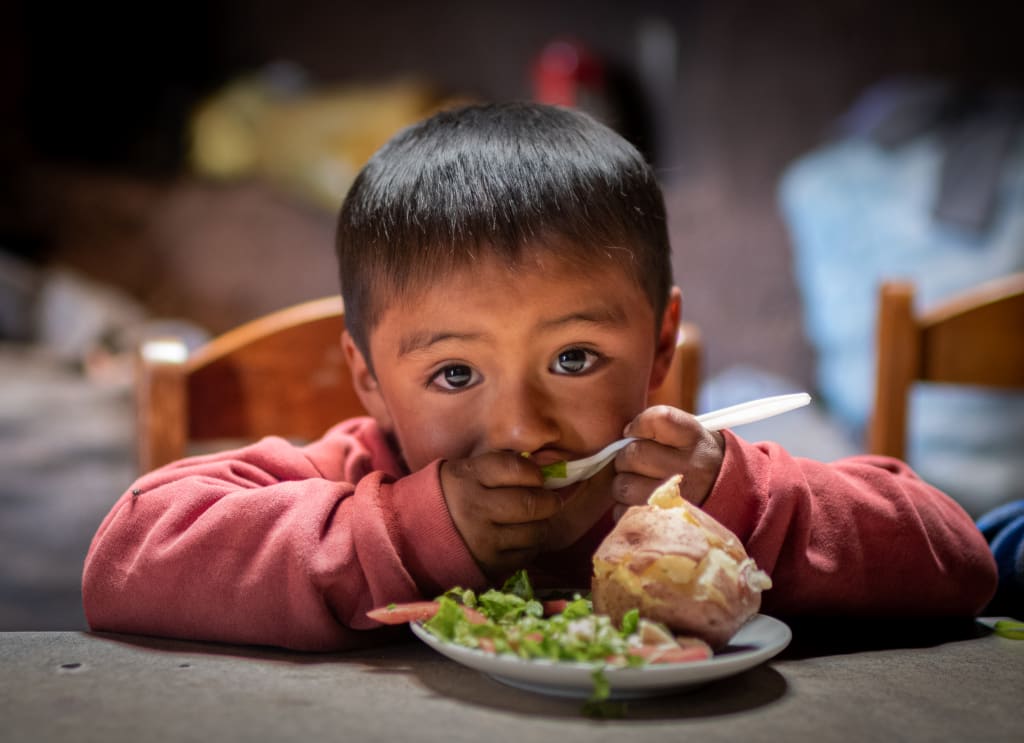 A little boy in red sits at the table with a plate of food and holds a fork to his mouth.
