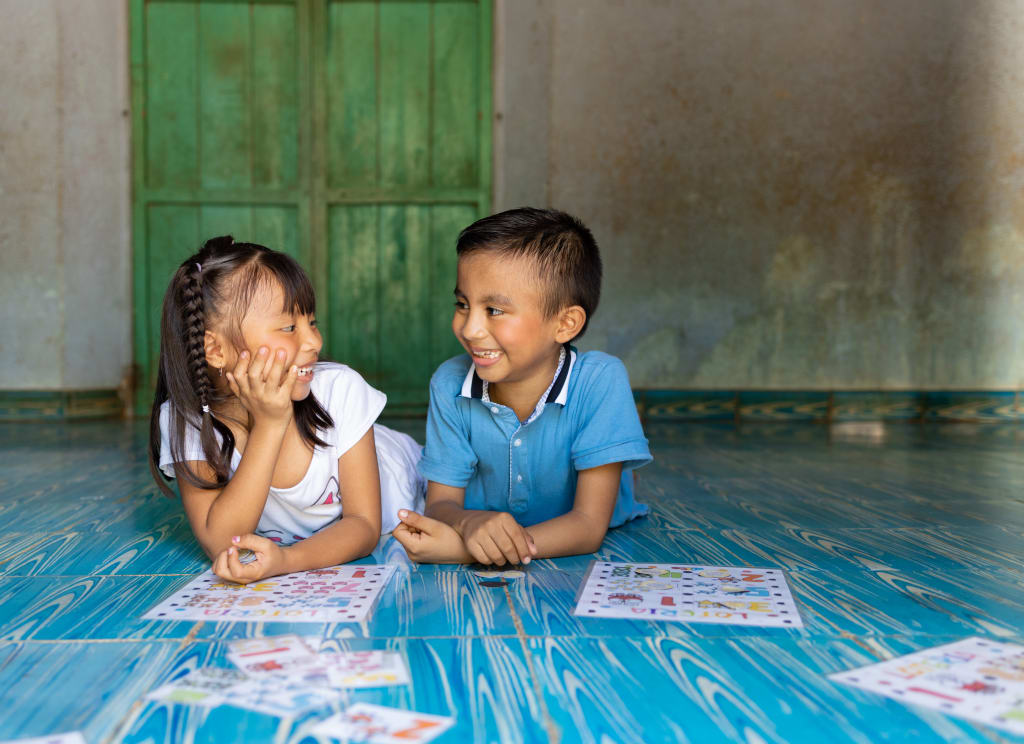 A boy in blue and a girl in white lay on their stomachs on a blue floor and smile at each other.