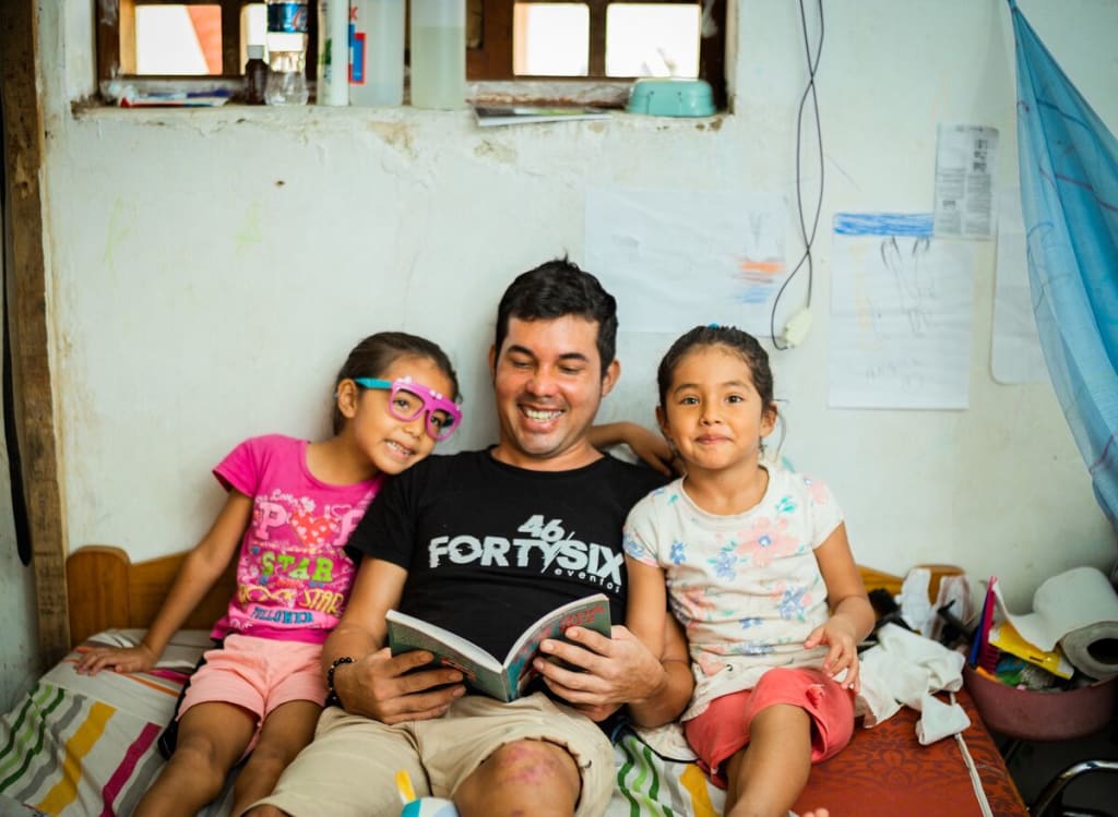 A dad in a black shirt sits between his two daughters and reads a book. They are all smiling.