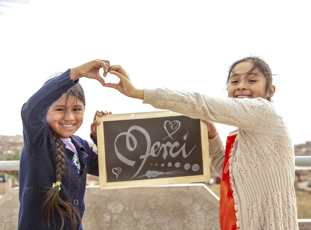 Two Bolivian girls hold up a chalkboard with the word "Merci" written on it.