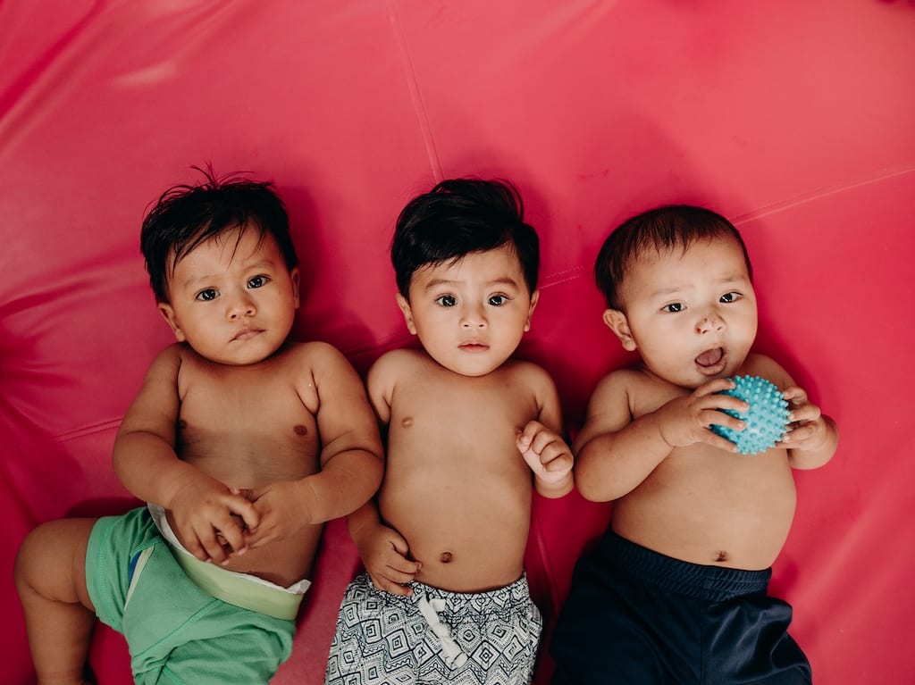 Three little baby boys lie on a red mat looking at the camera. They are children who are known, loved and protected, and loved by Jesus.