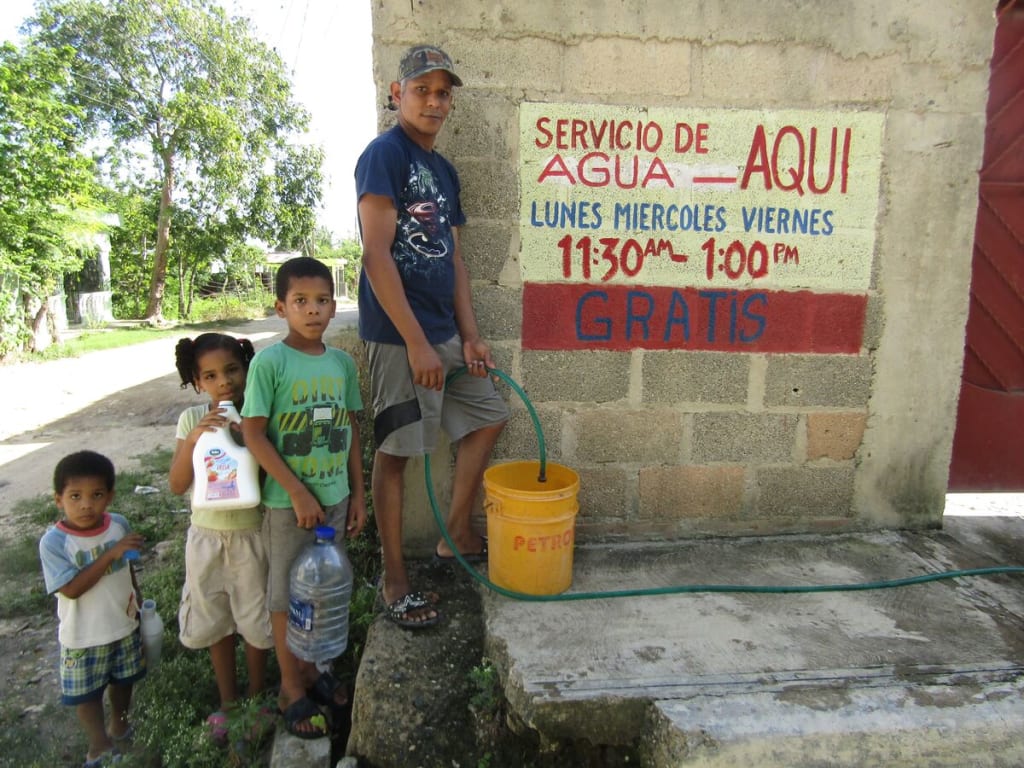 A man in a blue shirt and grey shorts stands with his children filling a yellow bucket with a hose.