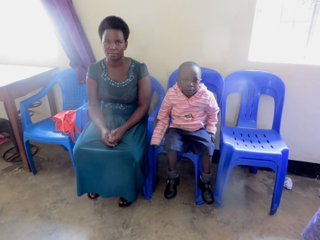 Saviour and his mom wait in his Compassion centre. They sit on blue chairs.