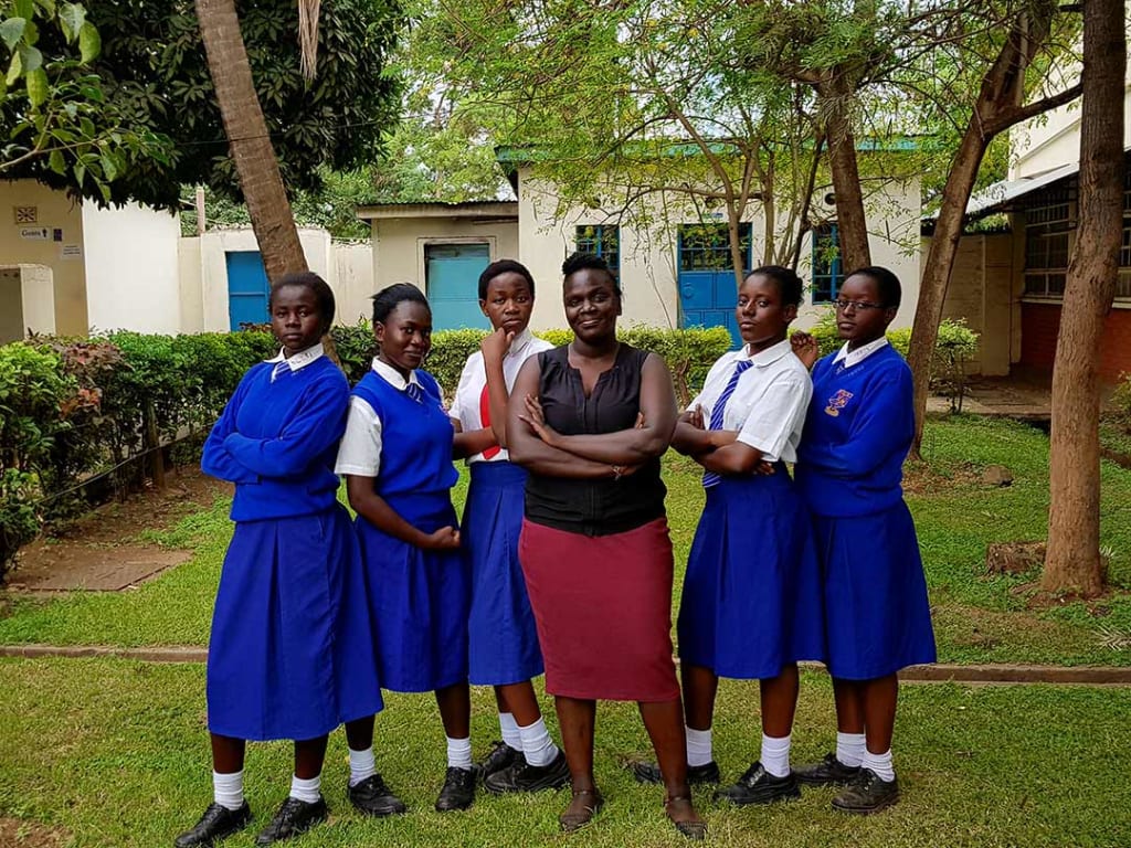 Six young women stand in the yard of their school. Five of them where blue and white school uniforms. One wears a black blouse and red dress.